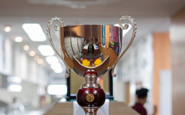 A large silver trophy cup with ornate handles and a dark red/maroon stem is displayed on a glass surface in what appears to be a military or institutional building. The trophy bears a plaque that references the 'Captain Edward F. Ney Memorial Award' and mentions 'Commander, Fleet Activities Yokosuka, Japan.' The background shows a blurred corridor with people and bulletin boards.
