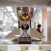 A large silver trophy cup with ornate handles and a dark red/maroon stem is displayed on a glass surface in what appears to be a military or institutional building. The trophy bears a plaque that references the 'Captain Edward F. Ney Memorial Award' and mentions 'Commander, Fleet Activities Yokosuka, Japan.' The background shows a blurred corridor with people and bulletin boards.