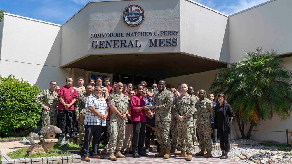 A group of U.S. Navy personnel and civilian staff pose for a formal group photo in front of the Commodore Matthew C. Perry General Mess facility, which bears a CFAY (Commander, Fleet Activities Yokosuka) emblem. Two individuals in the center appear to be exchanging or presenting an award or plaque. The setting appears to be a U.S. military installation, likely in Japan based on the CFAY insignia and surrounding vegetation.