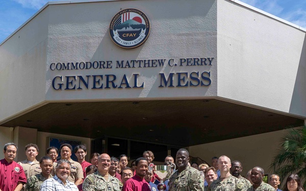 A group of U.S. Navy personnel and civilian staff pose for a formal group photo in front of the Commodore Matthew C. Perry General Mess facility, which bears a CFAY (Commander, Fleet Activities Yokosuka) emblem. Two individuals in the center appear to be exchanging or presenting an award or plaque. The setting appears to be a U.S. military installation, likely in Japan based on the CFAY insignia and surrounding vegetation.