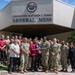 A group of U.S. Navy personnel and civilian staff pose for a formal group photo in front of the Commodore Matthew C. Perry General Mess facility, which bears a CFAY (Commander, Fleet Activities Yokosuka) emblem. Two individuals in the center appear to be exchanging or presenting an award or plaque. The setting appears to be a U.S. military installation, likely in Japan based on the CFAY insignia and surrounding vegetation.