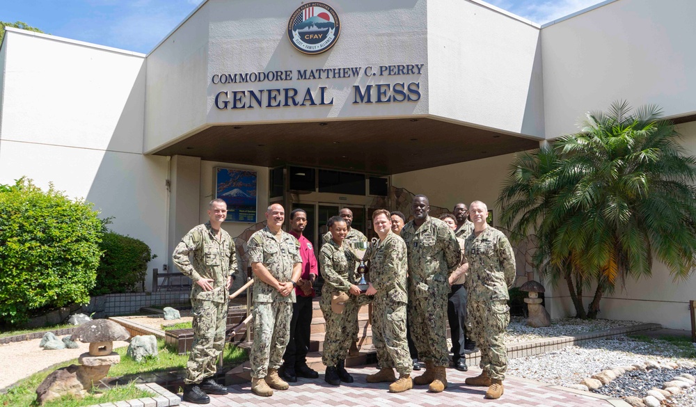 A group of approximately ten U.S. Navy personnel in camouflage uniforms are posed in front of the Commodore Matthew C. Perry General Mess building, which bears the CFAY (Commander, Fleet Activities Yokosuka) seal. Two individuals in the center are holding what appears to be a trophy or award cup, suggesting a recognition or award ceremony. The setting appears to be a U.S. naval installation, likely in Japan based on the CFAY insignia and surrounding landscape.
