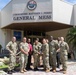 A group of approximately ten U.S. Navy personnel in camouflage uniforms are posed in front of the Commodore Matthew C. Perry General Mess building, which bears the CFAY (Commander, Fleet Activities Yokosuka) seal. Two individuals in the center are holding what appears to be a trophy or award cup, suggesting a recognition or award ceremony. The setting appears to be a U.S. naval installation, likely in Japan based on the CFAY insignia and surrounding landscape.