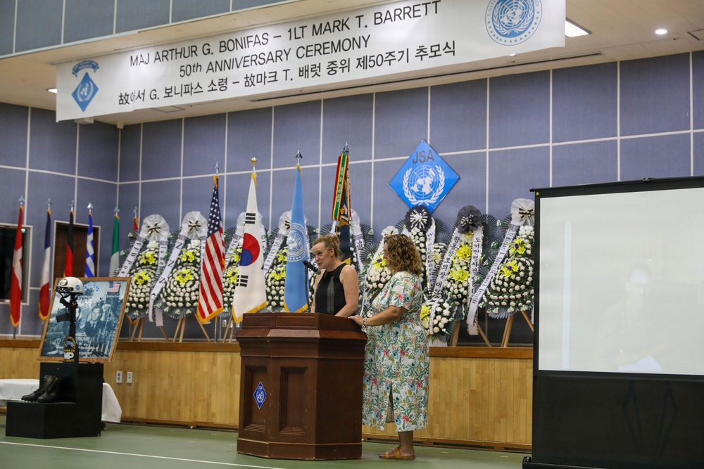 This image shows the 50th Anniversary Ceremony honoring Maj Arthur G. Bonifas and 1LT Mark T. Barrett, held at what appears to be a Joint Security Area (JSA) facility. Two women are standing at a wooden podium, likely delivering remarks during the memorial ceremony. The stage is decorated with numerous floral wreaths, international flags including the US, South Korean, and UN flags, and a JSA emblem.