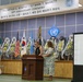 This image shows the 50th Anniversary Ceremony honoring Maj Arthur G. Bonifas and 1LT Mark T. Barrett, held at what appears to be a Joint Security Area (JSA) facility. Two women are standing at a wooden podium, likely delivering remarks during the memorial ceremony. The stage is decorated with numerous floral wreaths, international flags including the US, South Korean, and UN flags, and a JSA emblem.