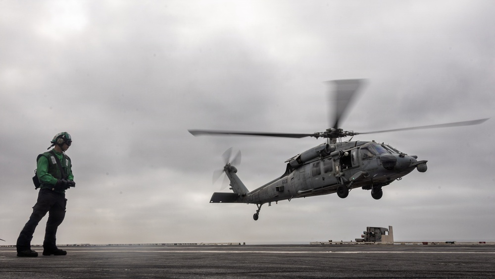 A military helicopter, appearing to be an MH-60 Seahawk, is hovering or landing on what appears to be a naval vessel flight deck. A flight deck crew member wearing a green jersey and helmet stands to the left, observing the aircraft. The scene takes place under overcast, grey skies suggesting maritime or coastal conditions.