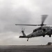 A military helicopter, appearing to be an MH-60 Seahawk, is hovering or landing on what appears to be a naval vessel flight deck. A flight deck crew member wearing a green jersey and helmet stands to the left, observing the aircraft. The scene takes place under overcast, grey skies suggesting maritime or coastal conditions.