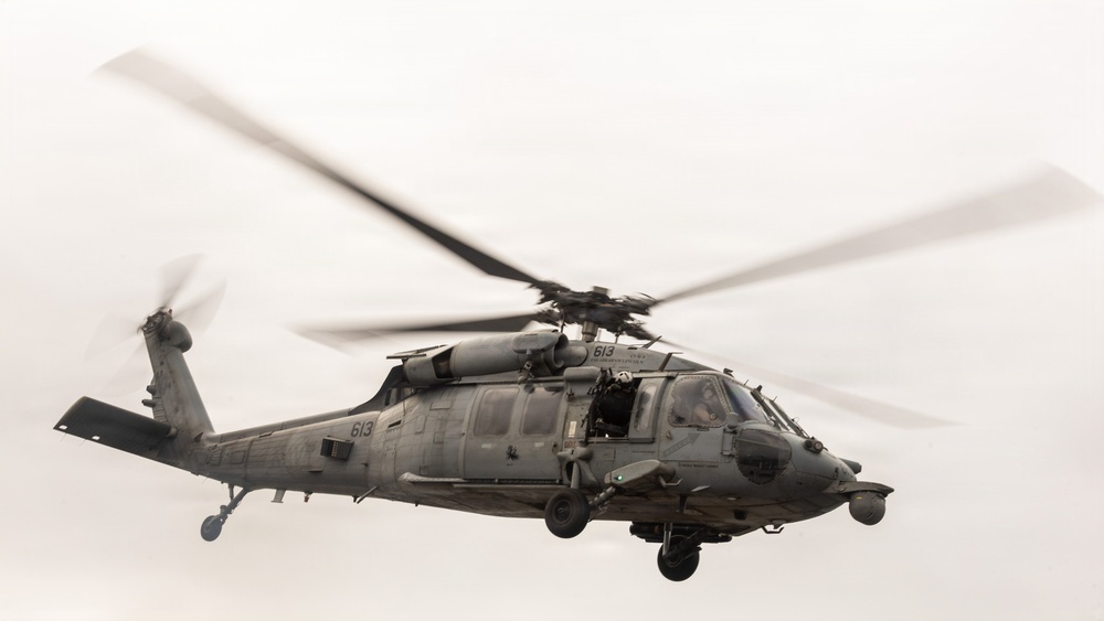 A military helicopter, identifiable as a Sikorsky HH-60 Pave Hawk or similar Black Hawk variant, is captured in flight against an overcast sky. The aircraft bears the tail number '613' visible on both the fuselage and tail boom, and is painted in a flat gray military finish. The rotor blades show significant motion blur, indicating active flight at the time of capture.