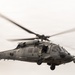 A military helicopter, identifiable as a Sikorsky HH-60 Pave Hawk or similar Black Hawk variant, is captured in flight against an overcast sky. The aircraft bears the tail number '613' visible on both the fuselage and tail boom, and is painted in a flat gray military finish. The rotor blades show significant motion blur, indicating active flight at the time of capture.
