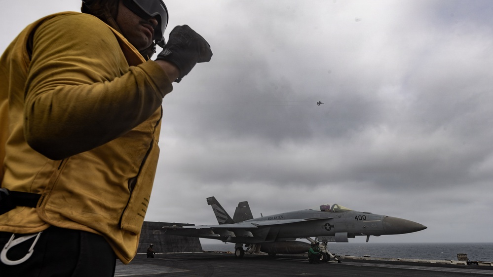 A U.S. Navy flight deck crew member in a yellow jersey stands on an aircraft carrier flight deck, observing operations. A fighter jet marked '400' — identifiable as an F/A-18 Hornet or Super Hornet — is positioned on the deck, while another aircraft is visible airborne in the overcast sky. The scene depicts active carrier flight operations at sea.