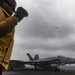 A U.S. Navy flight deck crew member in a yellow jersey stands on an aircraft carrier flight deck, observing operations. A fighter jet marked '400' — identifiable as an F/A-18 Hornet or Super Hornet — is positioned on the deck, while another aircraft is visible airborne in the overcast sky. The scene depicts active carrier flight operations at sea.