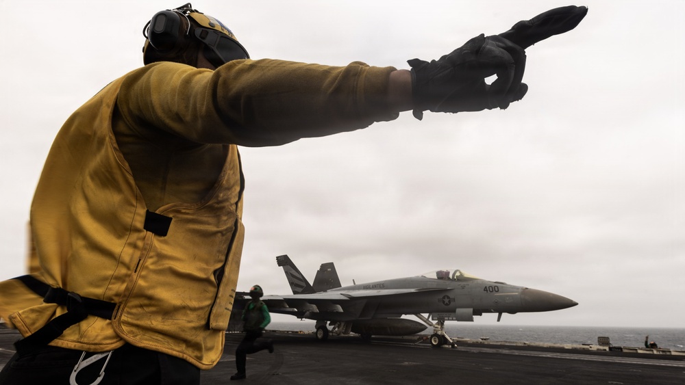A U.S. Navy flight deck crew member wearing a yellow jersey and black gloves is directing aircraft operations on an aircraft carrier flight deck. In the background, a U.S. Navy F/A-18 Super Hornet (modex 400) is positioned on the deck, likely preparing for launch or taxi. The overcast sky and open ocean are visible in the background, indicating the carrier is at sea.