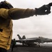 A U.S. Navy flight deck crew member wearing a yellow jersey and black gloves is directing aircraft operations on an aircraft carrier flight deck. In the background, a U.S. Navy F/A-18 Super Hornet (modex 400) is positioned on the deck, likely preparing for launch or taxi. The overcast sky and open ocean are visible in the background, indicating the carrier is at sea.
