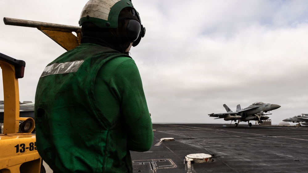 A flight deck crew member wearing a green jersey and helmet stands on an aircraft carrier flight deck observing a military jet aircraft on the runway. The green jersey indicates the crew member is likely a catapult and arresting gear officer or aircraft handling crew. The scene depicts active flight deck operations aboard a naval vessel under overcast skies.
