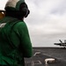 A flight deck crew member wearing a green jersey and helmet stands on an aircraft carrier flight deck observing a military jet aircraft on the runway. The green jersey indicates the crew member is likely a catapult and arresting gear officer or aircraft handling crew. The scene depicts active flight deck operations aboard a naval vessel under overcast skies.