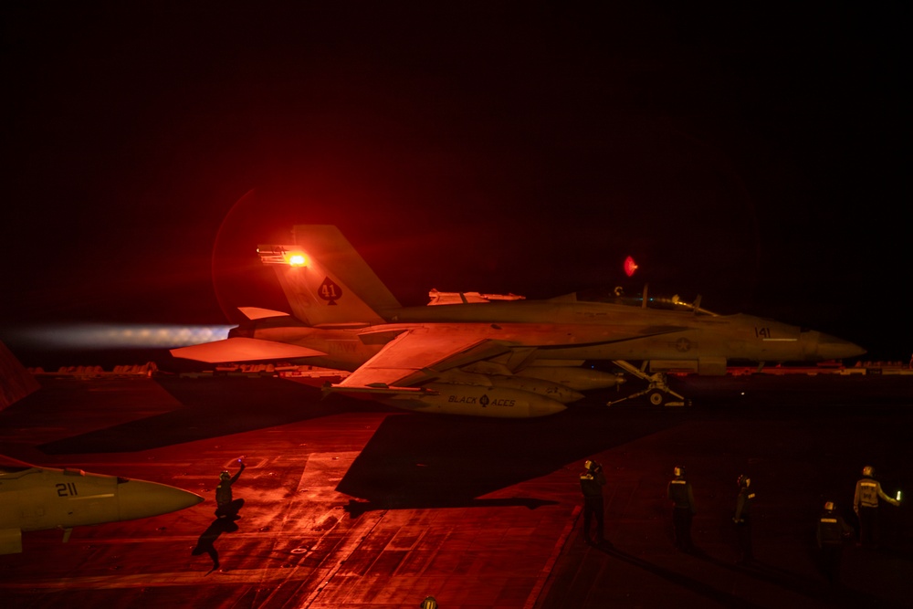 A U.S. Navy F/A-18 Super Hornet, marked 'Black Aces' with modex number 141, is photographed at night on an aircraft carrier flight deck with afterburners engaged, bathed in red lighting. The aircraft appears to be in a pre-launch or run-up position on the carrier deck. Ground crew personnel are visible in the background observing or directing flight deck operations.