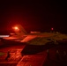 A U.S. Navy F/A-18 Super Hornet, marked 'Black Aces' with modex number 141, is photographed at night on an aircraft carrier flight deck with afterburners engaged, bathed in red lighting. The aircraft appears to be in a pre-launch or run-up position on the carrier deck. Ground crew personnel are visible in the background observing or directing flight deck operations.
