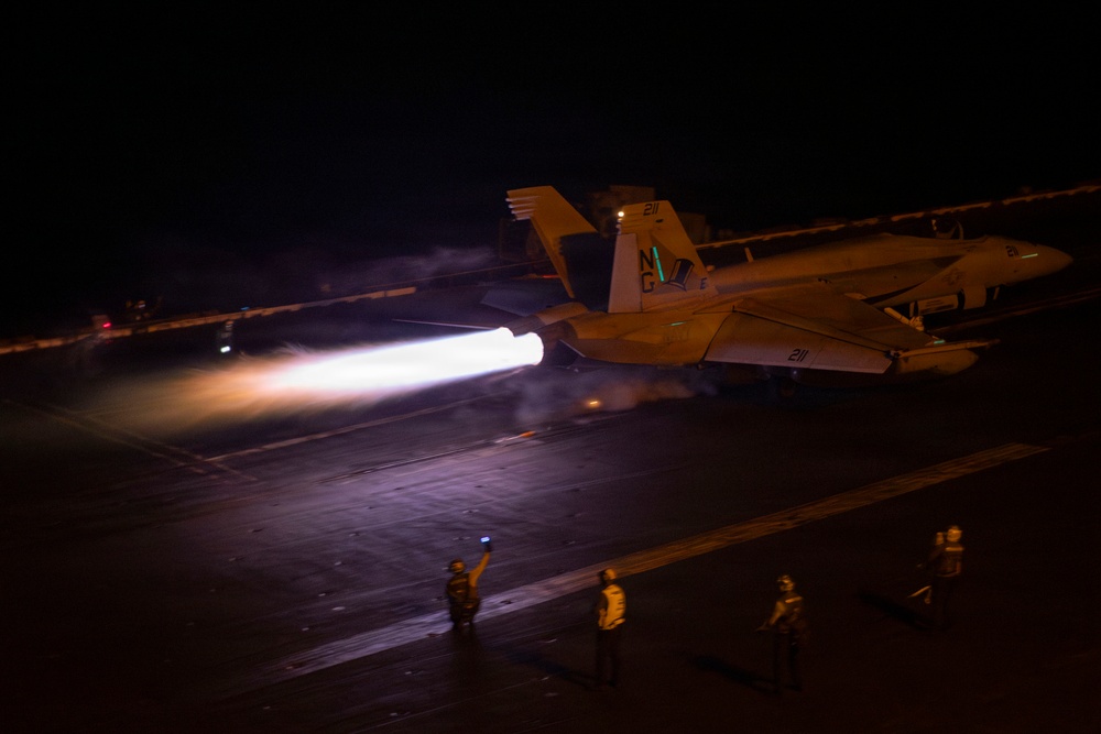 A military jet aircraft, marked with the number '211' and tail code 'NG', is launching from an aircraft carrier deck at night with full afterburner engaged, producing a bright white-blue exhaust plume. Multiple flight deck crew members in colored jerseys are visible on the deck, with one appearing to signal with a lighted wand. A second aircraft is visible parked on the deck in the background.