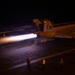 A military jet aircraft, marked with the number '211' and tail code 'NG', is launching from an aircraft carrier deck at night with full afterburner engaged, producing a bright white-blue exhaust plume. Multiple flight deck crew members in colored jerseys are visible on the deck, with one appearing to signal with a lighted wand. A second aircraft is visible parked on the deck in the background.