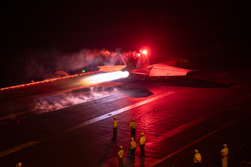 A military jet aircraft, marked with the number '211', is being catapult-launched from an aircraft carrier deck during nighttime operations. The scene is dramatically lit by red warning lights and the intense white glow of the aircraft's afterburners, with steam from the catapult system visible along the deck. Several flight deck crew members in yellow jerseys observe the launch from a safe distance.