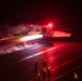 A military jet aircraft, marked with the number '211', is being catapult-launched from an aircraft carrier deck during nighttime operations. The scene is dramatically lit by red warning lights and the intense white glow of the aircraft's afterburners, with steam from the catapult system visible along the deck. Several flight deck crew members in yellow jerseys observe the launch from a safe distance.