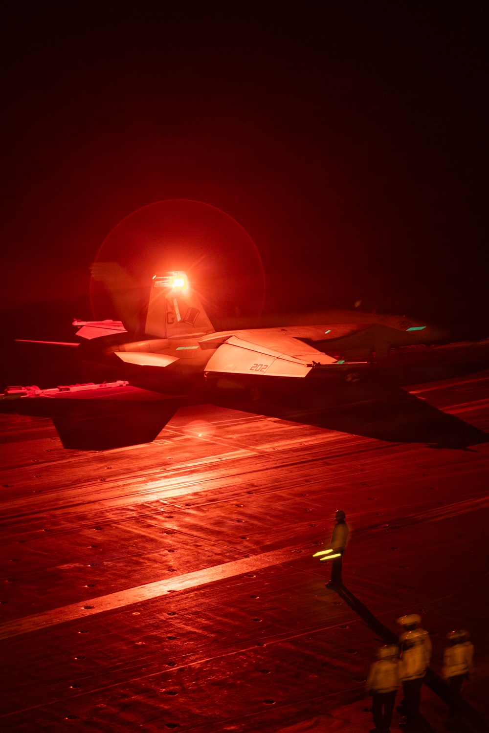 A military jet aircraft, marked with the number '202', is positioned on an aircraft carrier flight deck at night, bathed in intense red lighting. Deck crew personnel are visible in the foreground, wearing yellow jerseys with reflective markings. The scene depicts pre-launch or post-landing flight deck operations under nighttime conditions.