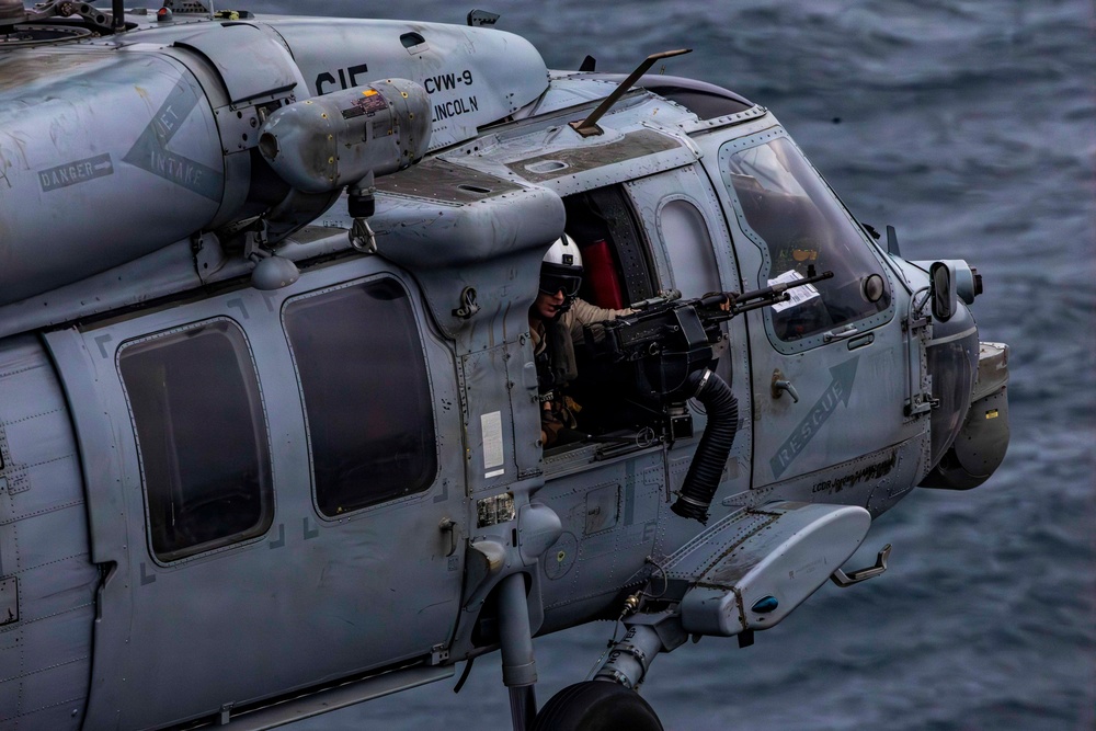 A U.S. Navy military helicopter, marked with 'CVW-9 LINCOLN' and tail code 'CIF', is flying over open water. A crew member wearing a flight helmet and visor is visible manning a door-mounted machine gun from the open side door. The word 'RESCUE' is stenciled on the fuselage, identifying this as likely an MH-60S or MH-60R Seahawk helicopter.