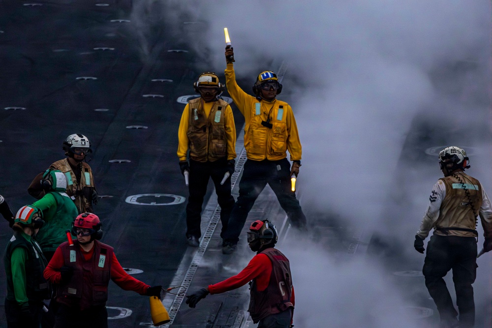 The image depicts flight deck operations aboard a U.S. Navy aircraft carrier, with crew members in color-coded jerseys and helmets performing their assigned roles. A central figure in a yellow jersey holds illuminated wands aloft, likely directing aircraft or signaling during launch or recovery operations. Steam or exhaust obscures part of the deck, adding to the dynamic and hazardous environment typical of carrier flight operations.