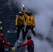 The image depicts flight deck operations aboard a U.S. Navy aircraft carrier, with crew members in color-coded jerseys and helmets performing their assigned roles. A central figure in a yellow jersey holds illuminated wands aloft, likely directing aircraft or signaling during launch or recovery operations. Steam or exhaust obscures part of the deck, adding to the dynamic and hazardous environment typical of carrier flight operations.