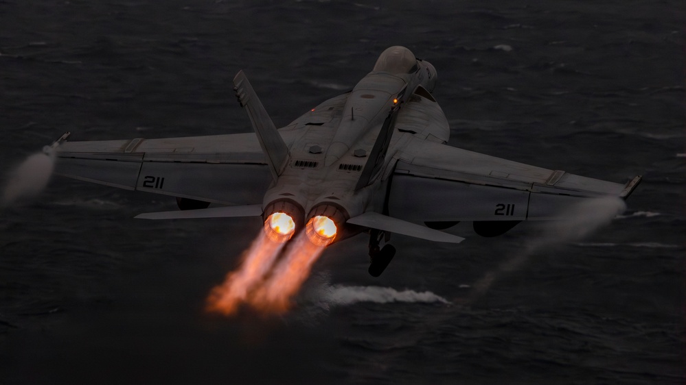 A carrier-based twin-engine jet fighter, marked with the number 211 on both wings, is captured launching at low altitude over open ocean in low-light conditions. Both engines are visibly in full afterburner, producing bright orange flame plumes. The aircraft appears to be in a steep climb shortly after catapult launch from a naval vessel.