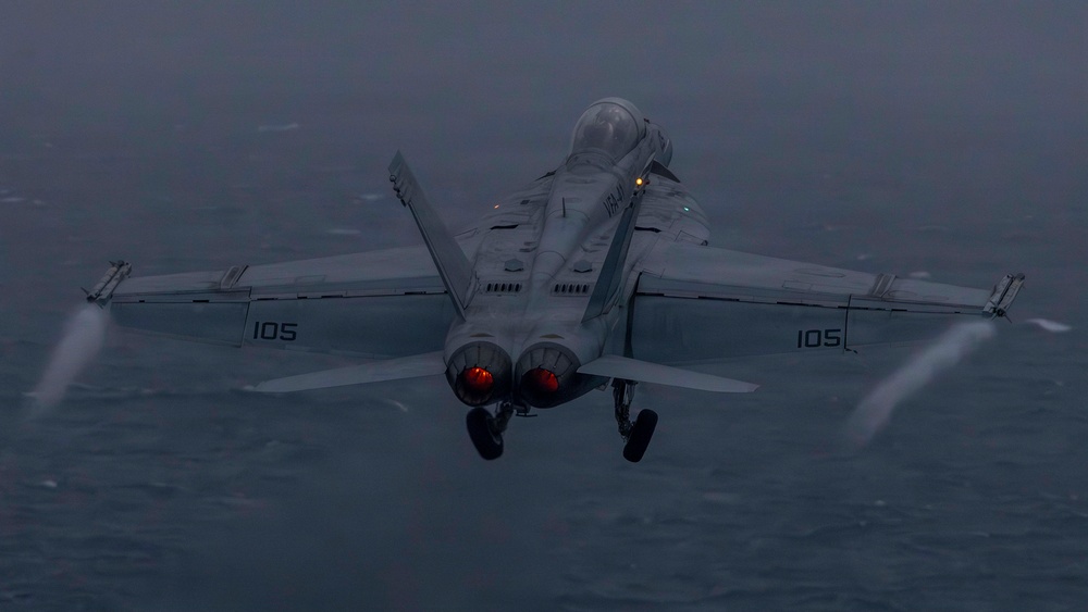 A U.S. Navy twin-engine carrier-based jet aircraft, marked with the number '105' on both wings, is photographed from a rear-facing perspective during low-light conditions over open water. The aircraft appears to be in a climb or just after launch, with landing gear still deployed and engine afterburners glowing red. Vapor condensation trails are visible along the wingtips, indicating high-speed, high-humidity flight conditions.
