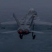 A U.S. Navy twin-engine carrier-based jet aircraft, marked with the number '105' on both wings, is photographed from a rear-facing perspective during low-light conditions over open water. The aircraft appears to be in a climb or just after launch, with landing gear still deployed and engine afterburners glowing red. Vapor condensation trails are visible along the wingtips, indicating high-speed, high-humidity flight conditions.