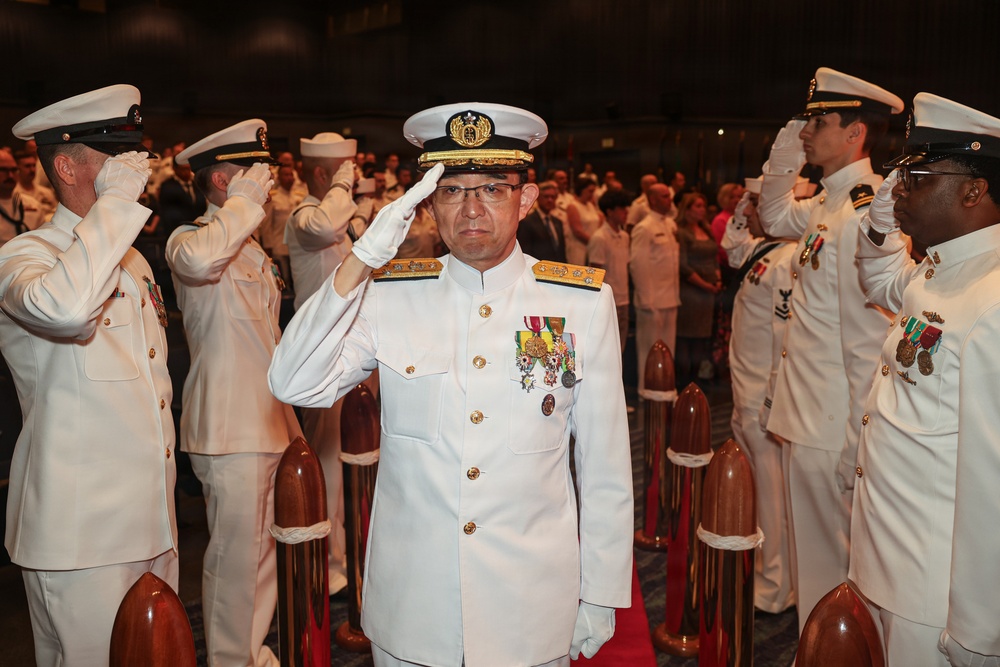A formal military ceremony featuring naval officers in white dress uniforms rendering salutes. The central figure, a senior officer adorned with multiple medals and gold-braided cap insignia, stands at attention on a red carpet flanked by wooden bollards. A large assembly of uniformed personnel and civilians is visible in the background.