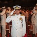 A formal military ceremony featuring naval officers in white dress uniforms rendering salutes. The central figure, a senior officer adorned with multiple medals and gold-braided cap insignia, stands at attention on a red carpet flanked by wooden bollards. A large assembly of uniformed personnel and civilians is visible in the background.