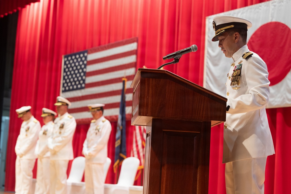 A U.S. Navy officer in white dress uniform speaks at a wooden podium during a formal military ceremony. The backdrop features both the American flag and the Japanese flag hung against red curtains, suggesting a bilateral or joint military event. Several other naval officers in white dress uniforms stand at attention on the stage behind the speaker.