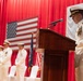 A U.S. Navy officer in white dress uniform speaks at a wooden podium during a formal military ceremony. The backdrop features both the American flag and the Japanese flag hung against red curtains, suggesting a bilateral or joint military event. Several other naval officers in white dress uniforms stand at attention on the stage behind the speaker.