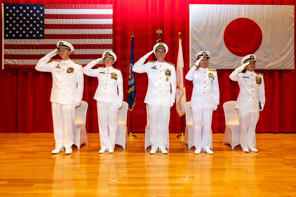 Five naval officers in white dress uniforms stand at salute on a wooden stage during a formal military ceremony. The backdrop features both the United States flag and the Japanese flag, along with color guard flags, indicating a joint US-Japan naval event. The formal setting and synchronized saluting suggest a change of command or official bilateral military ceremony.