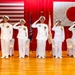 Five naval officers in white dress uniforms stand at salute on a wooden stage during a formal military ceremony. The backdrop features both the United States flag and the Japanese flag, along with color guard flags, indicating a joint US-Japan naval event. The formal setting and synchronized saluting suggest a change of command or official bilateral military ceremony.