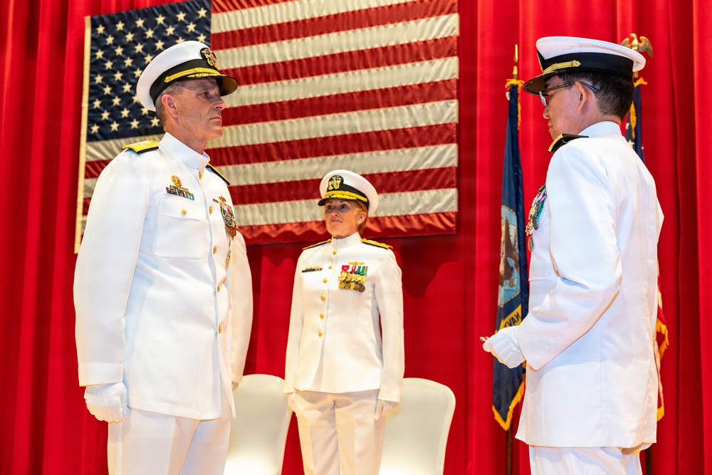 Three U.S. Navy officers in white dress uniforms are participating in a formal military ceremony, likely a change of command or promotion event. Two officers face each other in the foreground while a third stands at attention in the background. The setting features an American flag and a Navy organizational flag mounted on a red-draped stage.