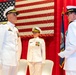 Three U.S. Navy officers in white dress uniforms are participating in a formal military ceremony, likely a change of command or promotion event. Two officers face each other in the foreground while a third stands at attention in the background. The setting features an American flag and a Navy organizational flag mounted on a red-draped stage.