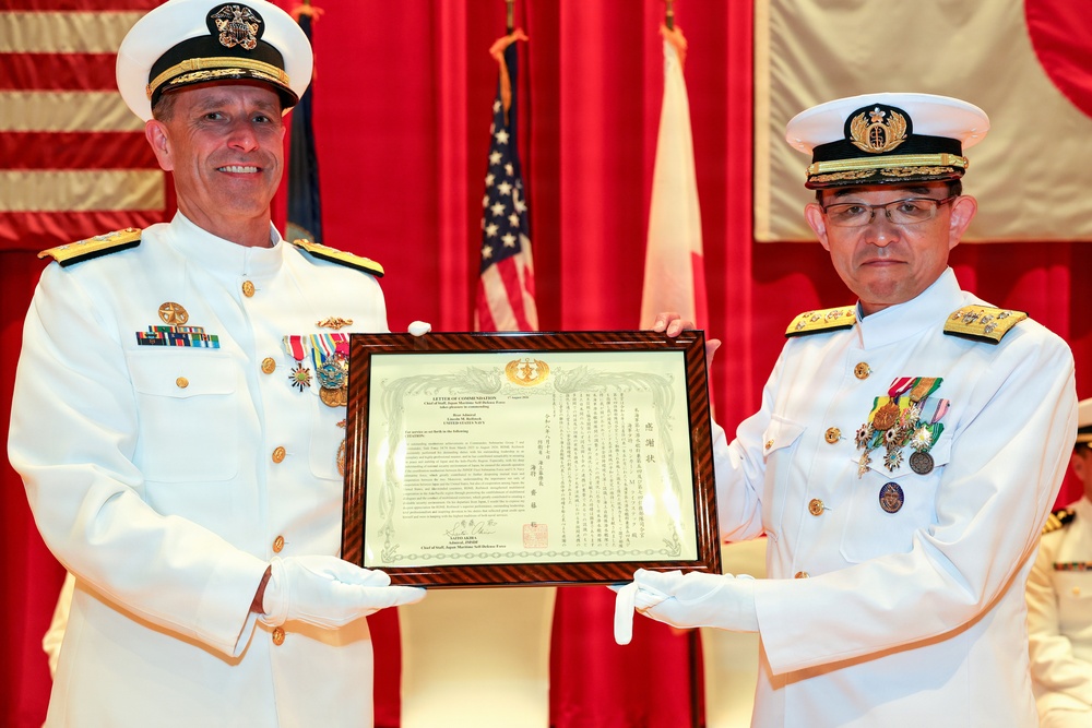 Two senior naval officers in white dress uniforms are jointly presenting or receiving a framed bilingual certificate or letter of commendation. The document appears to contain both English and Japanese text, suggesting a joint US-Japan military ceremony. Both officers are wearing multiple medals and decorations, indicating high rank.