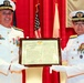 Two senior naval officers in white dress uniforms are jointly presenting or receiving a framed bilingual certificate or letter of commendation. The document appears to contain both English and Japanese text, suggesting a joint US-Japan military ceremony. Both officers are wearing multiple medals and decorations, indicating high rank.