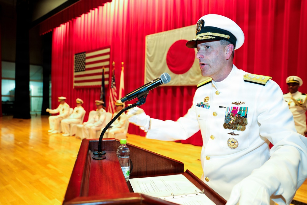 A senior U.S. Navy officer in white dress uniform is delivering a speech at a formal military ceremony, standing at a wooden podium with a microphone. The setting appears to be a joint U.S.-Japan military event, evidenced by both the American flag and Japanese flag (Hinomaru) displayed on a red-curtained stage. Several other naval personnel in white uniforms are seated in the background.