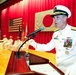 A senior U.S. Navy officer in white dress uniform is delivering a speech at a formal military ceremony, standing at a wooden podium with a microphone. The setting appears to be a joint U.S.-Japan military event, evidenced by both the American flag and Japanese flag (Hinomaru) displayed on a red-curtained stage. Several other naval personnel in white uniforms are seated in the background.