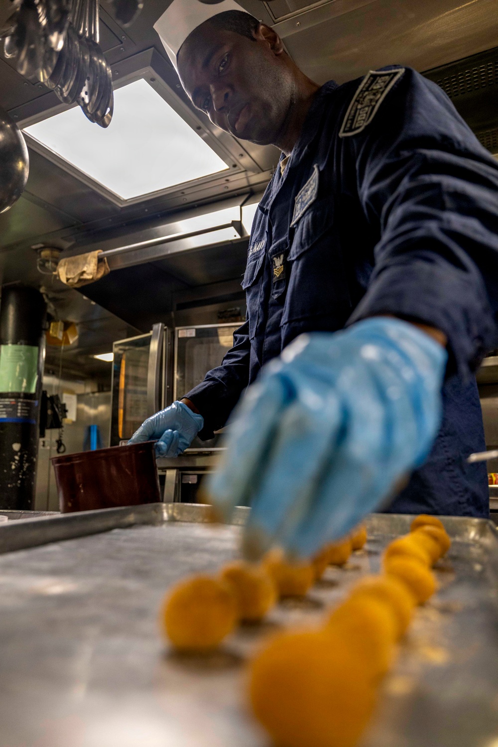 A U.S. Navy service member wearing a navy blue uniform and blue latex gloves is working in a shipboard galley (kitchen). The individual appears to be preparing food, arranging small round yellow food items on a metal tray. The low-angle shot provides a dramatic perspective of the kitchen environment aboard what appears to be a naval vessel.