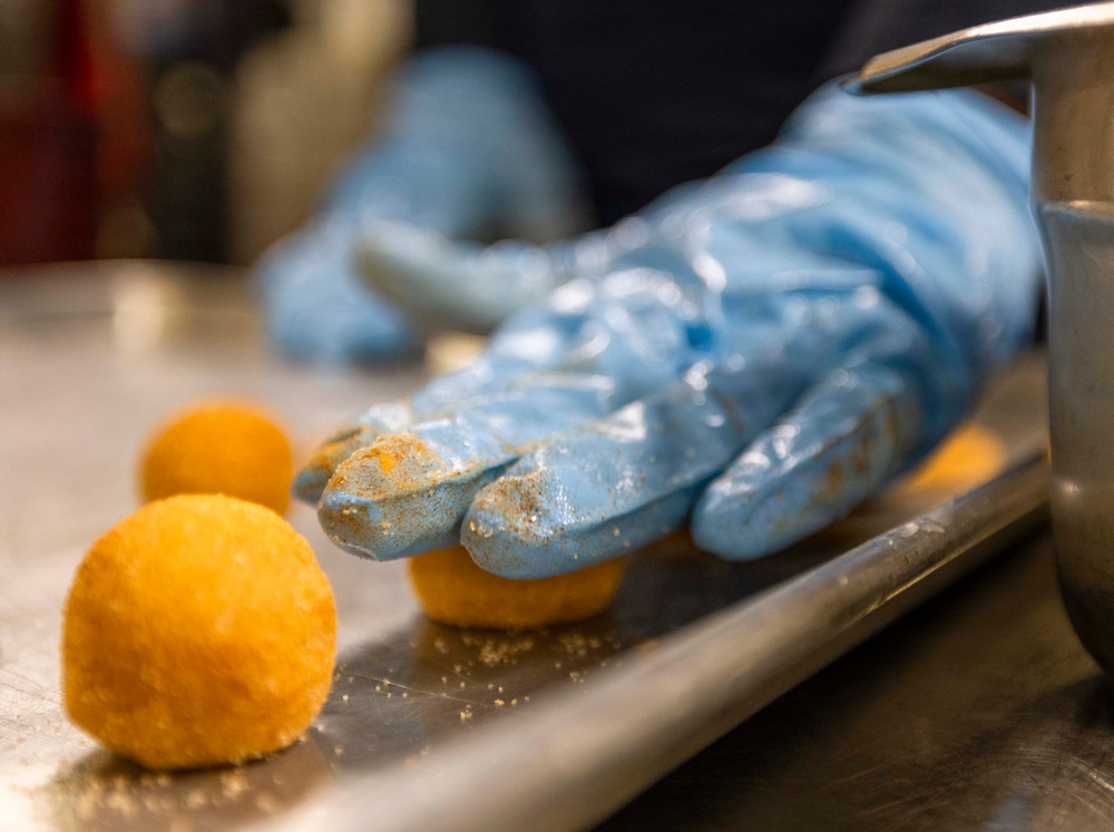 A food preparation scene showing a person wearing blue nitrile gloves handling round, breaded or coated food balls on a stainless steel tray. The gloves are visibly coated with orange/yellow residue from the food being shaped. The close-up perspective emphasizes the hands-on food production process in what appears to be a commercial kitchen.