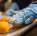 A food preparation scene showing a person wearing blue nitrile gloves handling round, breaded or coated food balls on a stainless steel tray. The gloves are visibly coated with orange/yellow residue from the food being shaped. The close-up perspective emphasizes the hands-on food production process in what appears to be a commercial kitchen.