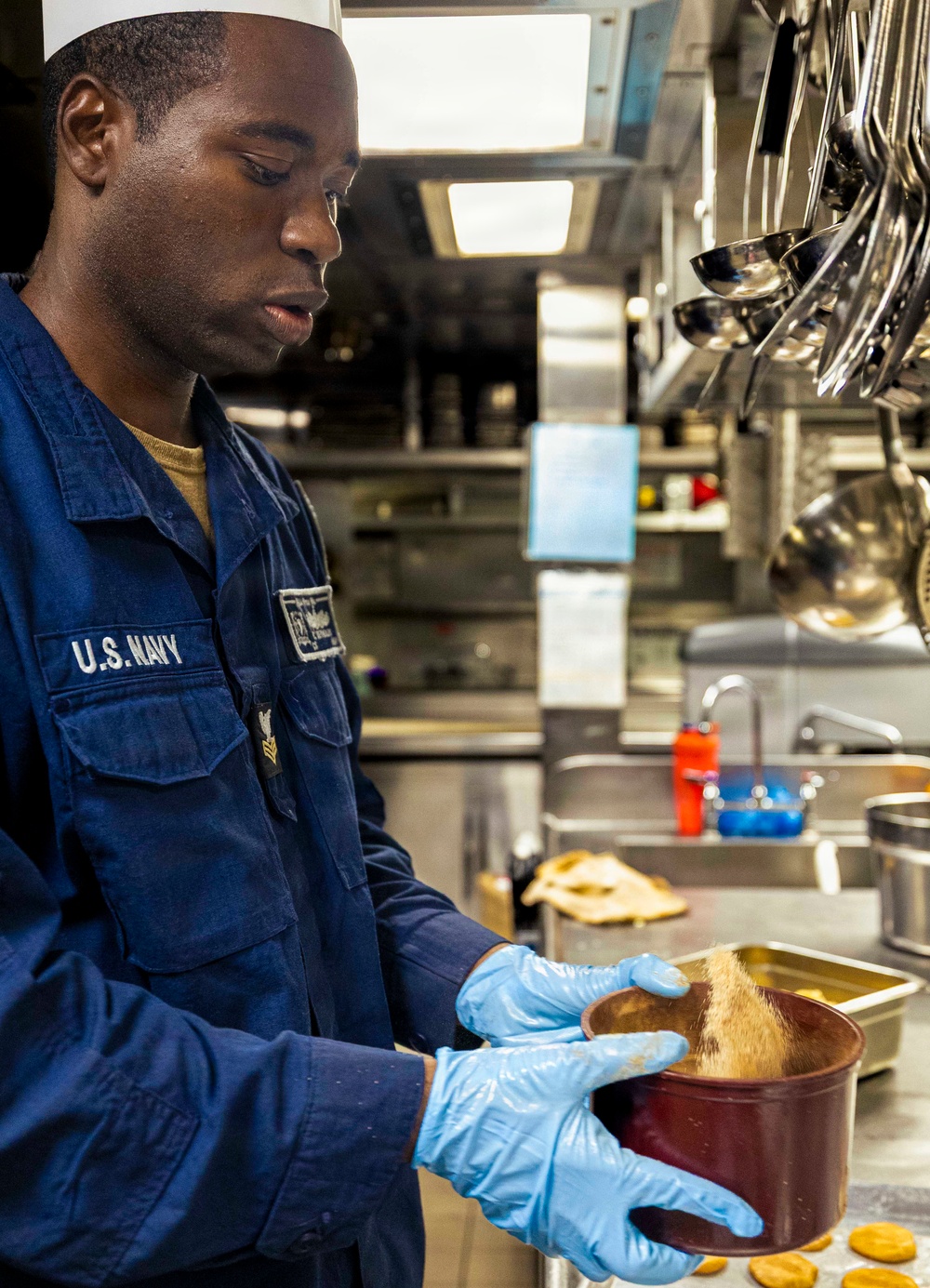 A U.S. Navy sailor in a ship's galley kitchen is preparing food while wearing blue latex gloves and a white cook's hat. The sailor is dressed in navy blue utility coveralls with 'U.S. NAVY' clearly visible on the chest. He is focused on pouring or mixing an ingredient into a round metal container.