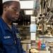 A U.S. Navy sailor in a ship's galley kitchen is preparing food while wearing blue latex gloves and a white cook's hat. The sailor is dressed in navy blue utility coveralls with 'U.S. NAVY' clearly visible on the chest. He is focused on pouring or mixing an ingredient into a round metal container.