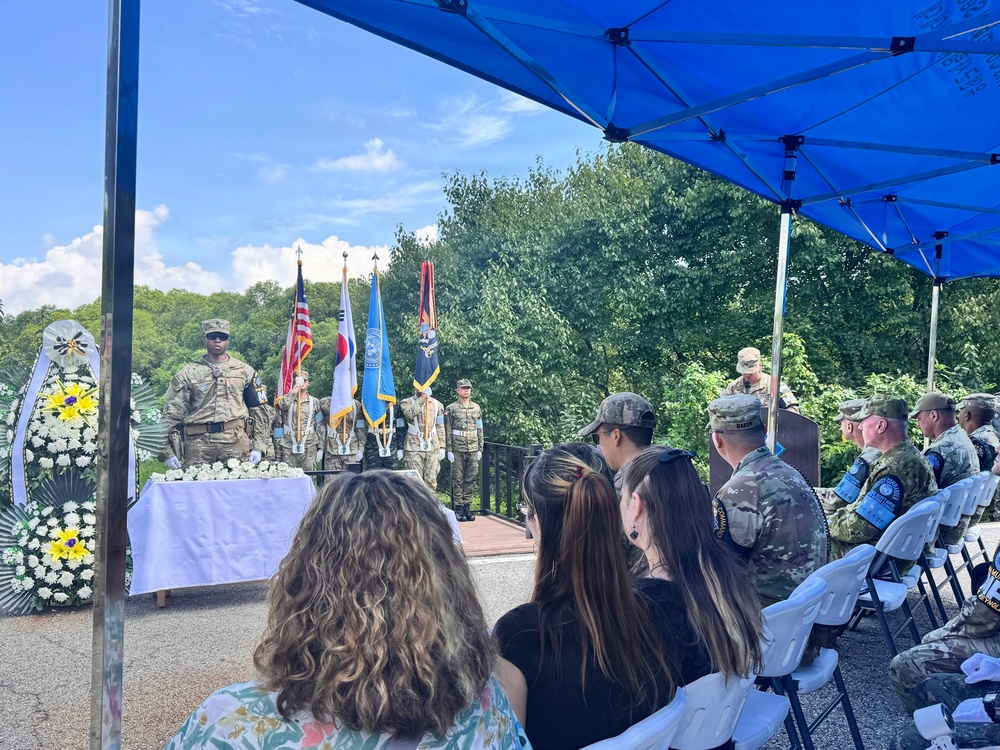 A military memorial or commemorative ceremony is taking place outdoors under a blue canopy tent. A color guard is presenting multiple flags including the American flag and what appears to be the South Korean flag, suggesting a joint US-Korean military event. Attendees including both military personnel in camouflage uniforms and civilians are seated facing a stage with a white-draped table adorned with floral wreaths.